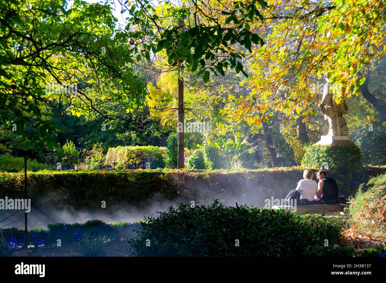 Twilight rays of the sun passing through the branches of autumnal trees next to two people sitting on their backs on a stone bench in the Royal Botani Stock Photo