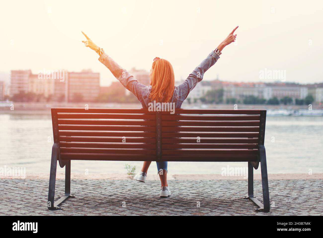Young redhead woman arms raised on bench in susnet at riverbank back view Stock Photo