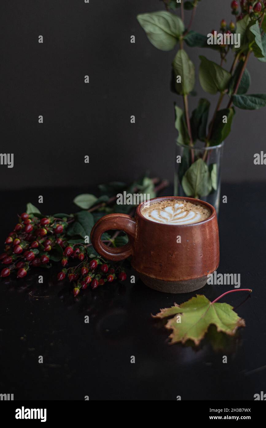 Maple spiced latte in handmade rust-colored ceramic mug with tulip latte art surrounded by fall leaf arrangement on black coffee counter. Stock Photo