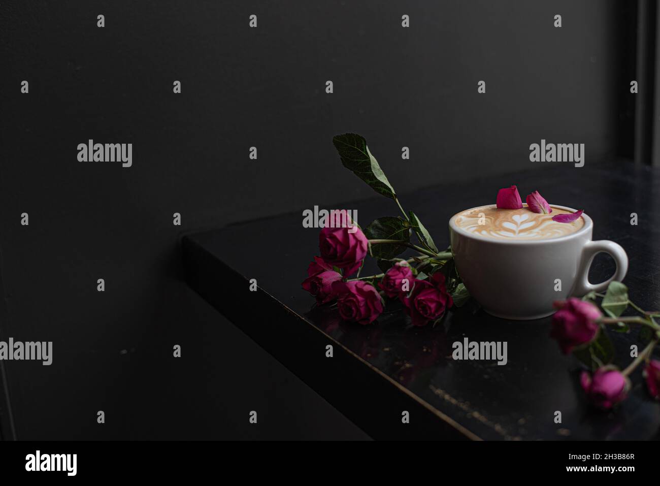Rose cardamom latte with latte art in white latte mug on dark coffee shop counter garnished with rose petals and styled with pink roses. Stock Photo