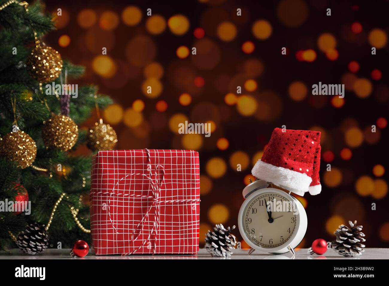 New Year's composition. Gift box and alarm clock in a Santa hat next to a decorated Christmas tree on a blurred background of lights. Stock Photo