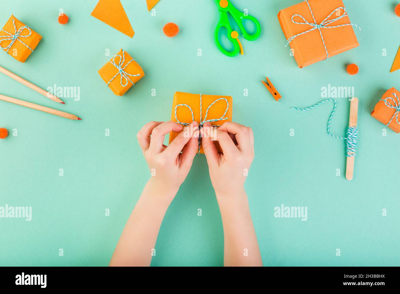 Child is whapping gift in orange paper on blue table top view. Season gift-giving concept. Made with his own hands. Stock Photo