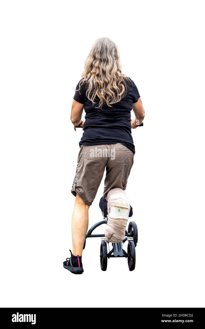 Back of injured woman in shorts with long hair with bandaged foot and knee scooter isolated on whilte. Stock Photo