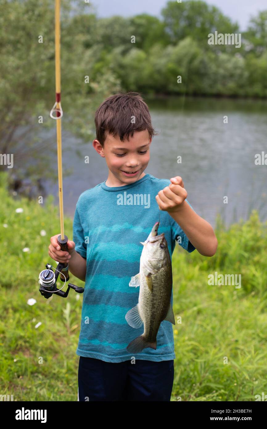 Cute little boy smiling as he learns to fish in a pond at an outdoor park.  Young black boy holding a fishing pole as he attempts to catch a fish Stock  Photo