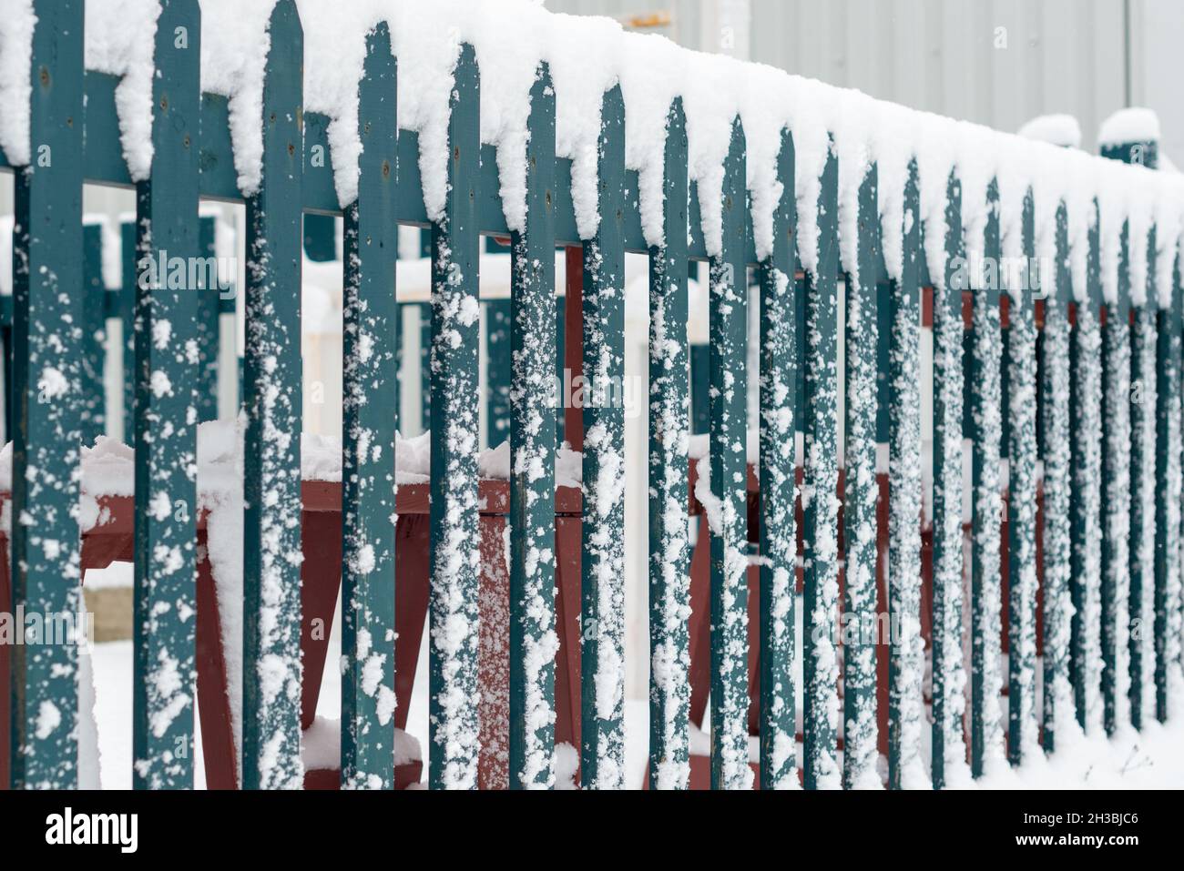 A dark green wood picket fence separating a road and field is covered in fresh white snow.  The ground is covered in fresh snow and large snowflakes. Stock Photo