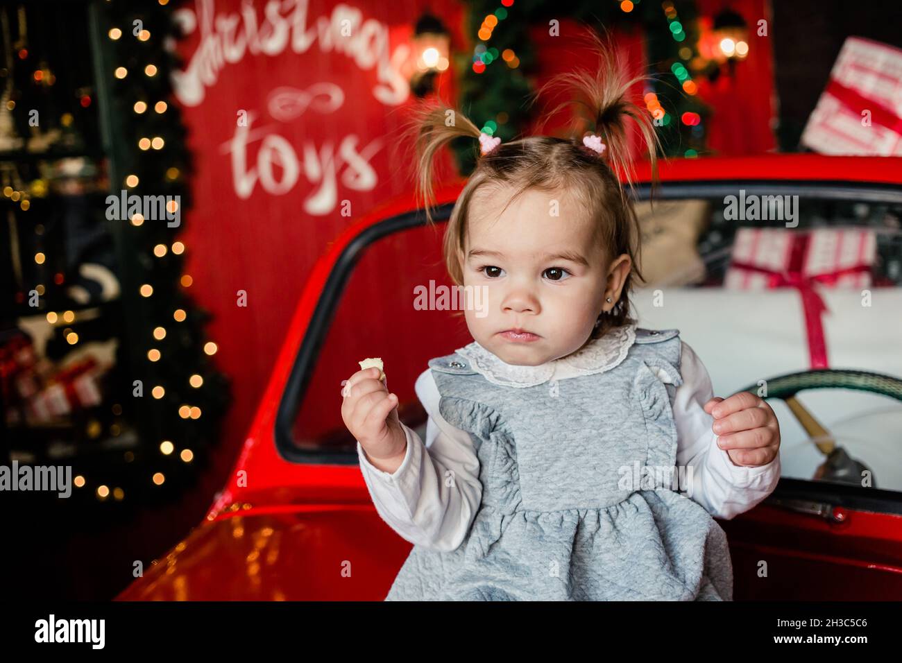 Joyful baby sitting on red Christmas car and eats christmas cookies in the living room at home. Copy space. Stock Photo