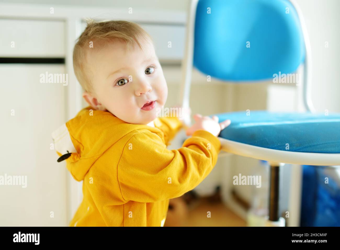 Adorable baby boy standing by the chair on bright living room. Toddler learning how to stand up unassisted. Stock Photo