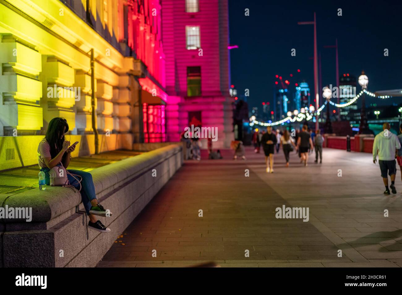 LONDON - SEPTEMBER 14, 2021: A young woman uses a smartphone while sat on a wall next to colorfully lit County Hall at night Stock Photo