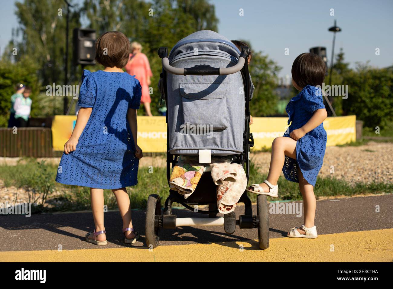 Two children next to the stroller on a walk on a sunny day. Girls in blue dresses, a view from the back. City holiday, celebration in the park Stock Photo