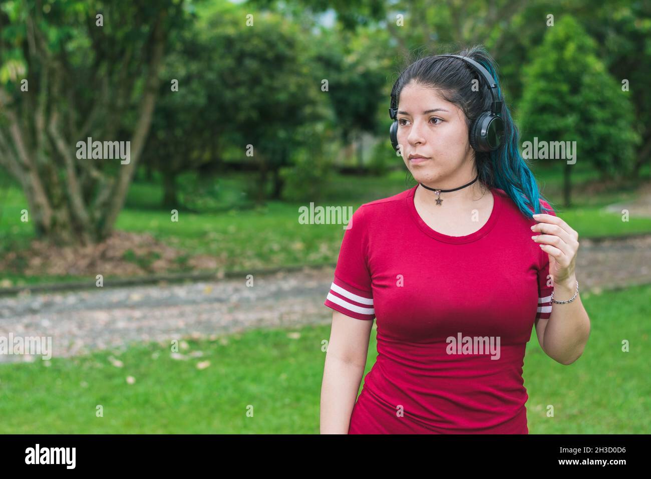 beautiful college woman with blue hair and red dress, listening to music with her wireless headphones in a park, stroking her blue hair while looking Stock Photo