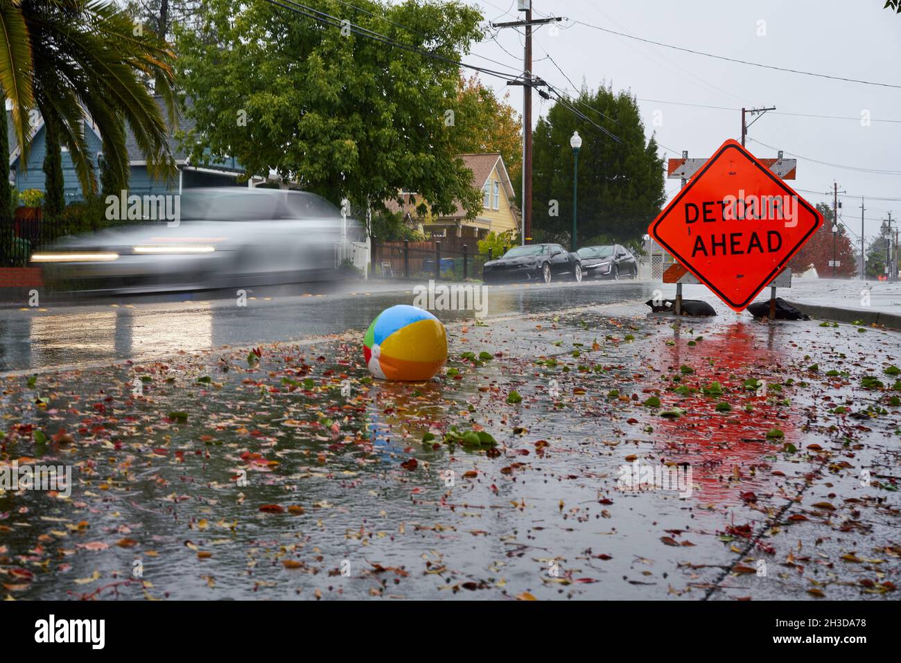 Car drives on wet street with a detour sign, fallen leaves and beach ball, in Windsor, California. Slow shutter shot with motion blur. Stock Photo