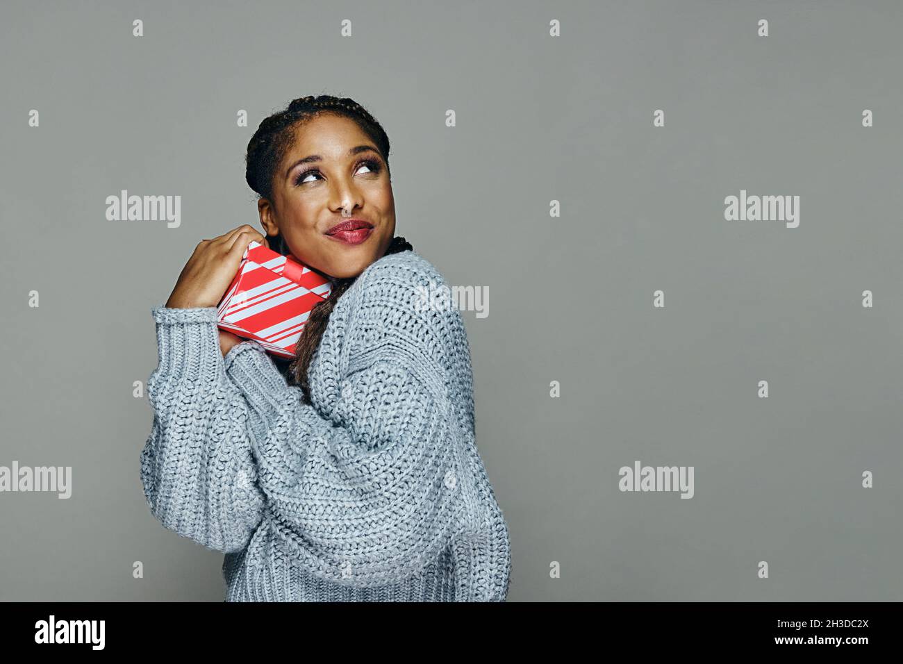 Happy attractive young woman in sweater hugging tight Christmas present while looking up against gray background Stock Photo