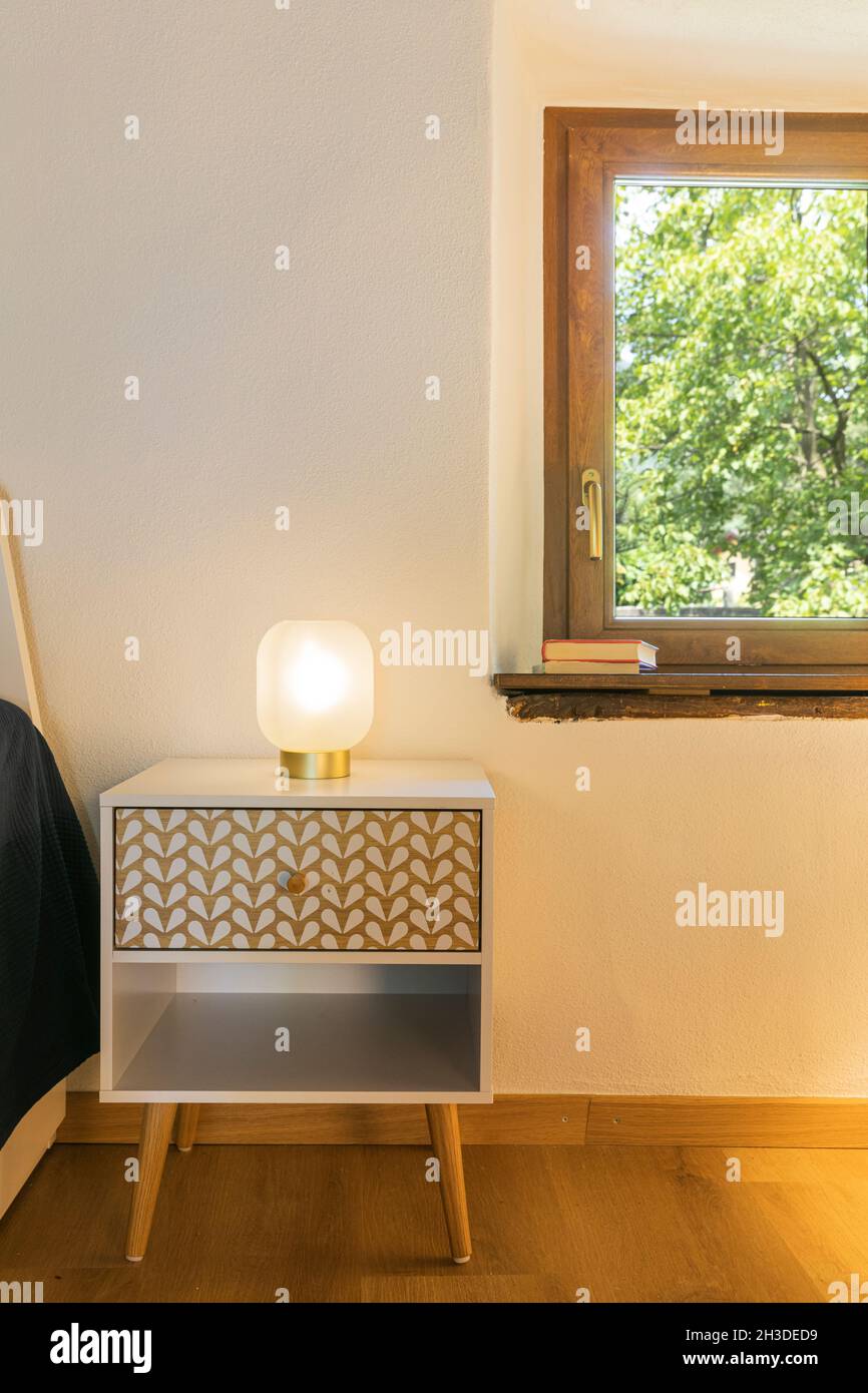 Detail of a bedroom, view of the bedside table with its light on. The wall is white and the view of the window overlooks the woods. Stock Photo