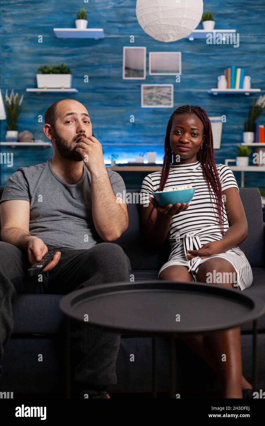 Interracial couple sitting on couch watching television at home. African american woman holding bowl with popcorn while caucasian man switching channels with TV remote control. Stock Photo