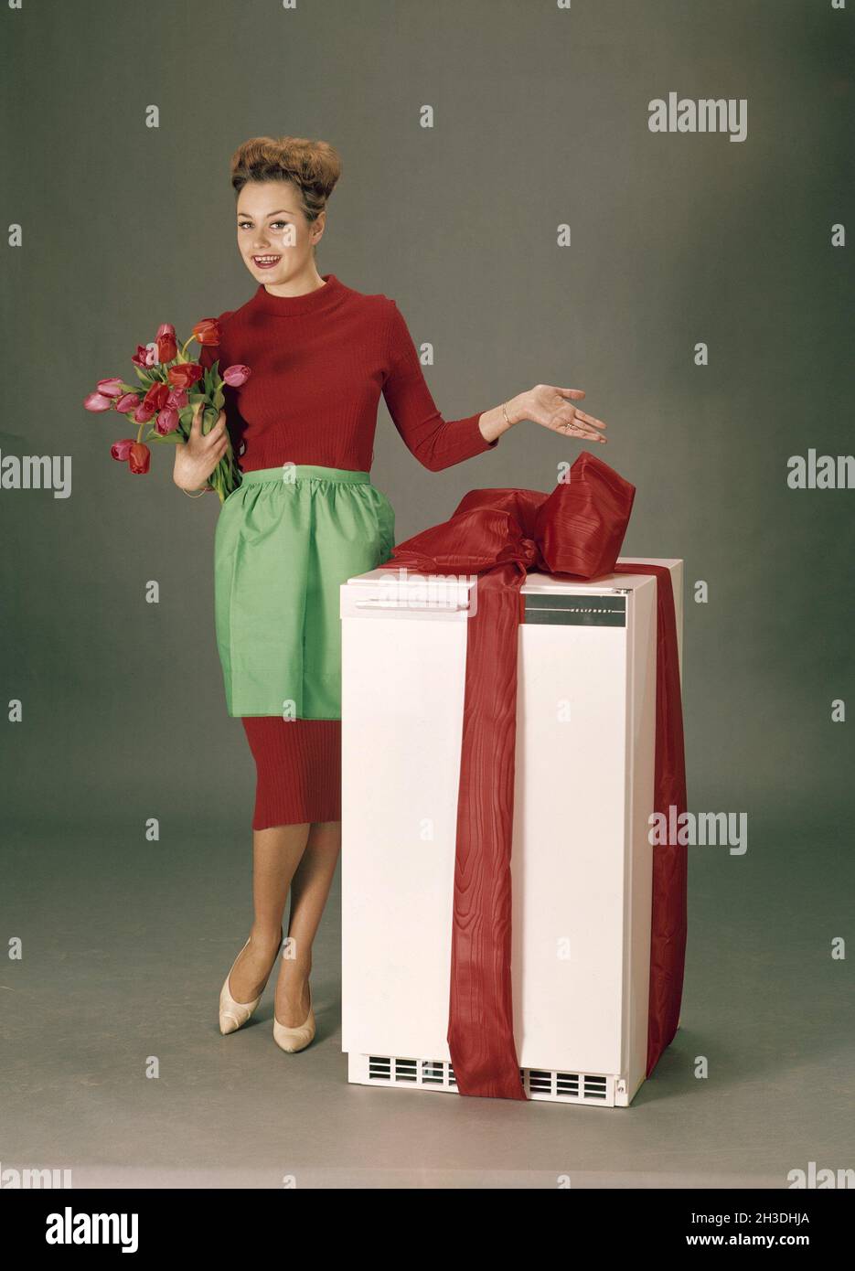In the 1960s. A woman in a red dress with a green apron is standing beside a refrigerator wrapped in a red ribbon as if she is presenting it as the first price in a competition. Sweden 1960s Stock Photo
