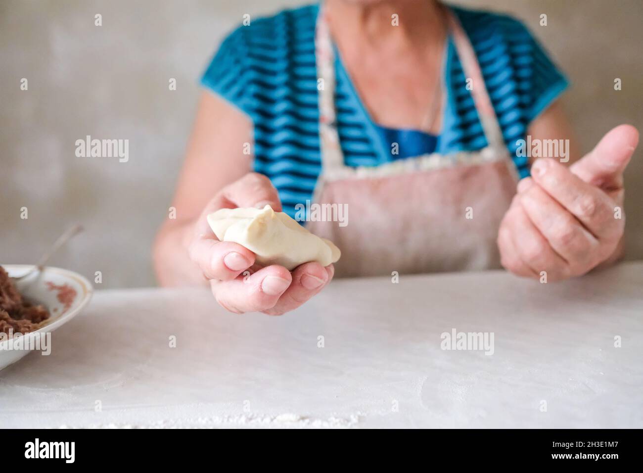 Grandmother holds in her hand a freshly molded manta. A dish of dough and minced meat Stock Photo