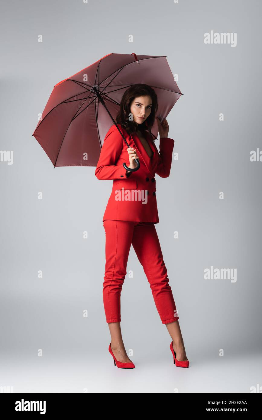 full length of young woman in red suit standing under umbrella on grey Stock Photo