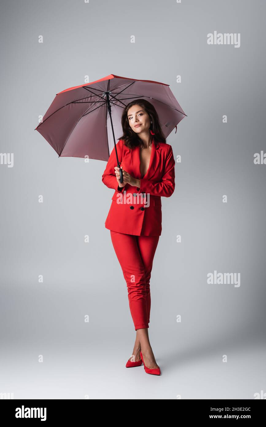 full length of brunette woman in red suit standing under umbrella on grey Stock Photo