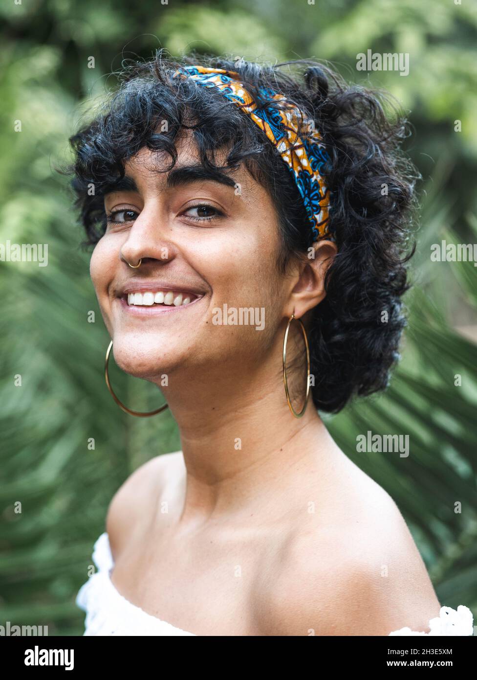 Cheerful charming ethnic female in headband on curly hair looking at camera  in green summer garden Stock Photo - Alamy