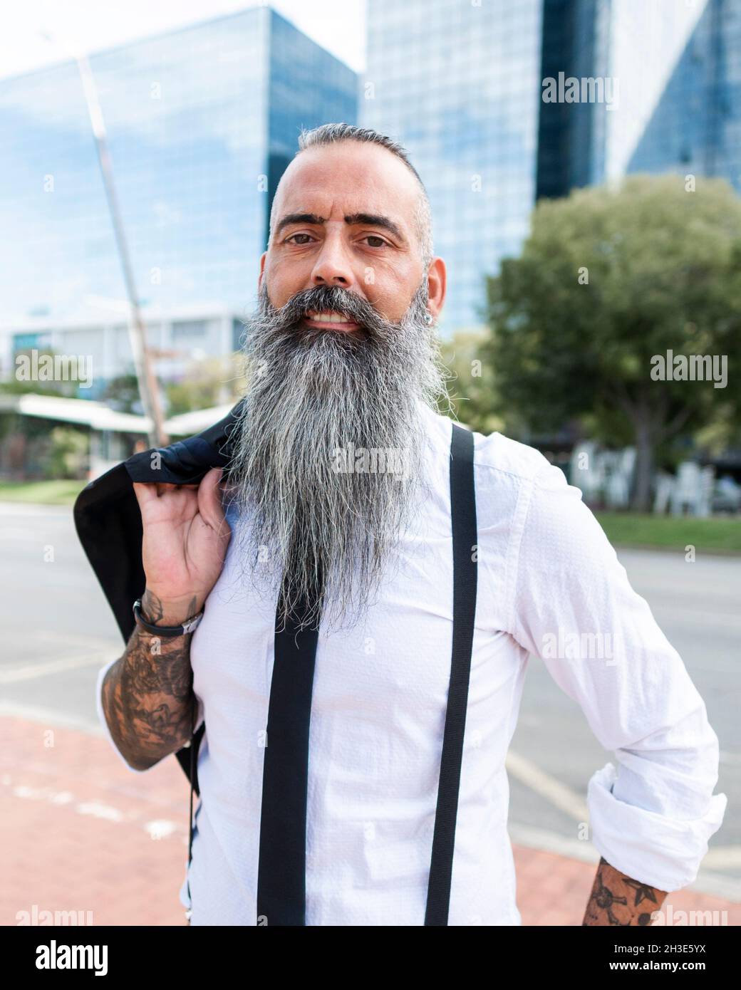 Confident bearded male with tattoo wearing stylish outfit looking at camera while standing on street with modern buildings in city Stock Photo