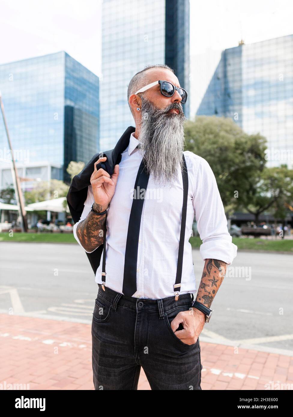 Confident bearded male with tattoo in sunglasses wearing stylish outfit looking away while standing on street with modern buildings in city Stock Photo