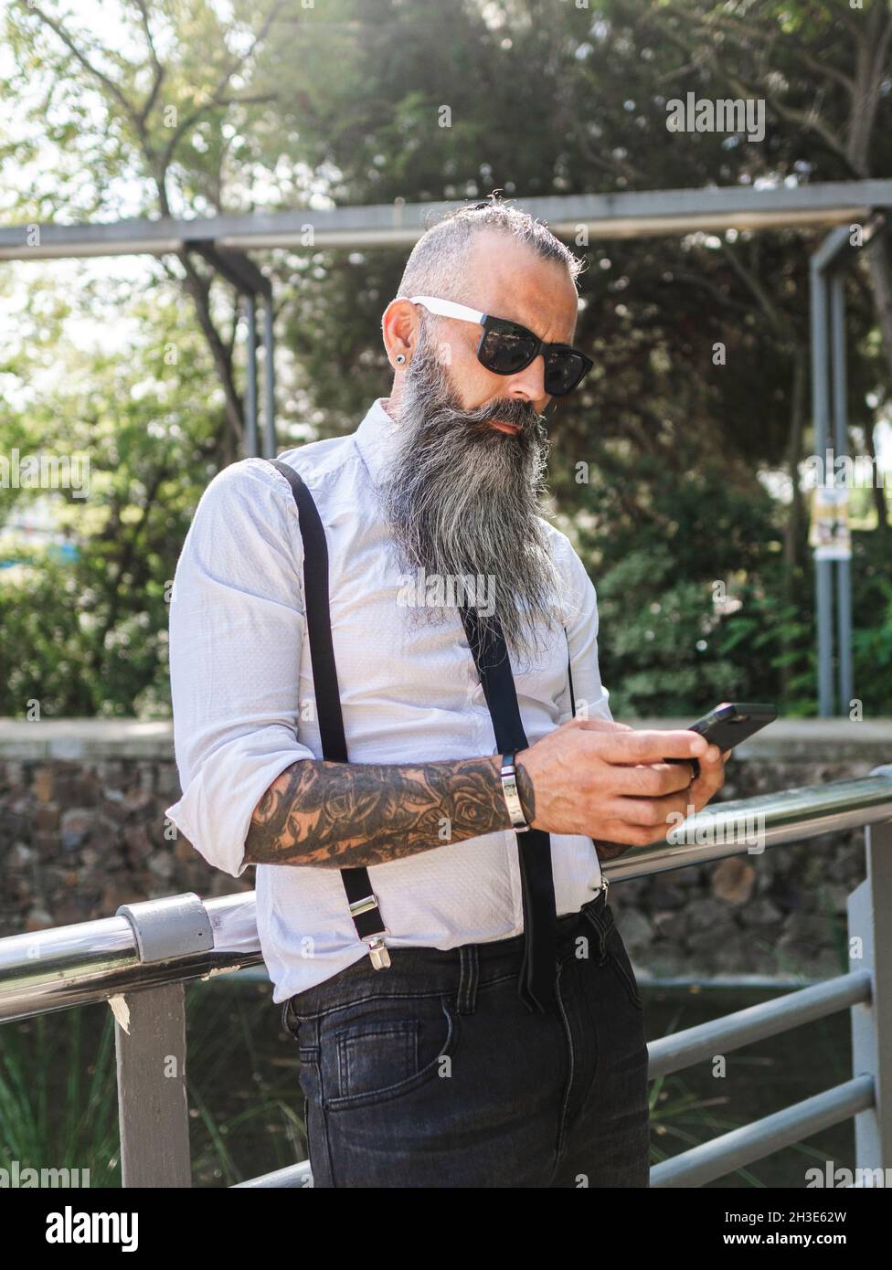 Confident bearded hipster in stylish outfit and sunglasses text messaging on cellphone while standing near metal fence on sunny street Stock Photo