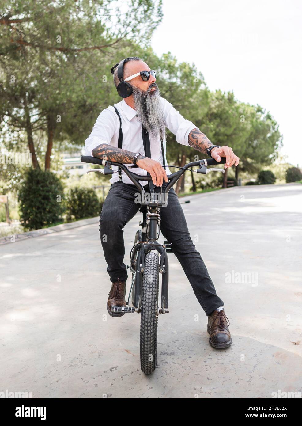 Confident male hipster in white shirt listening to music in headphones while sitting on bicycle on street with green trees Stock Photo