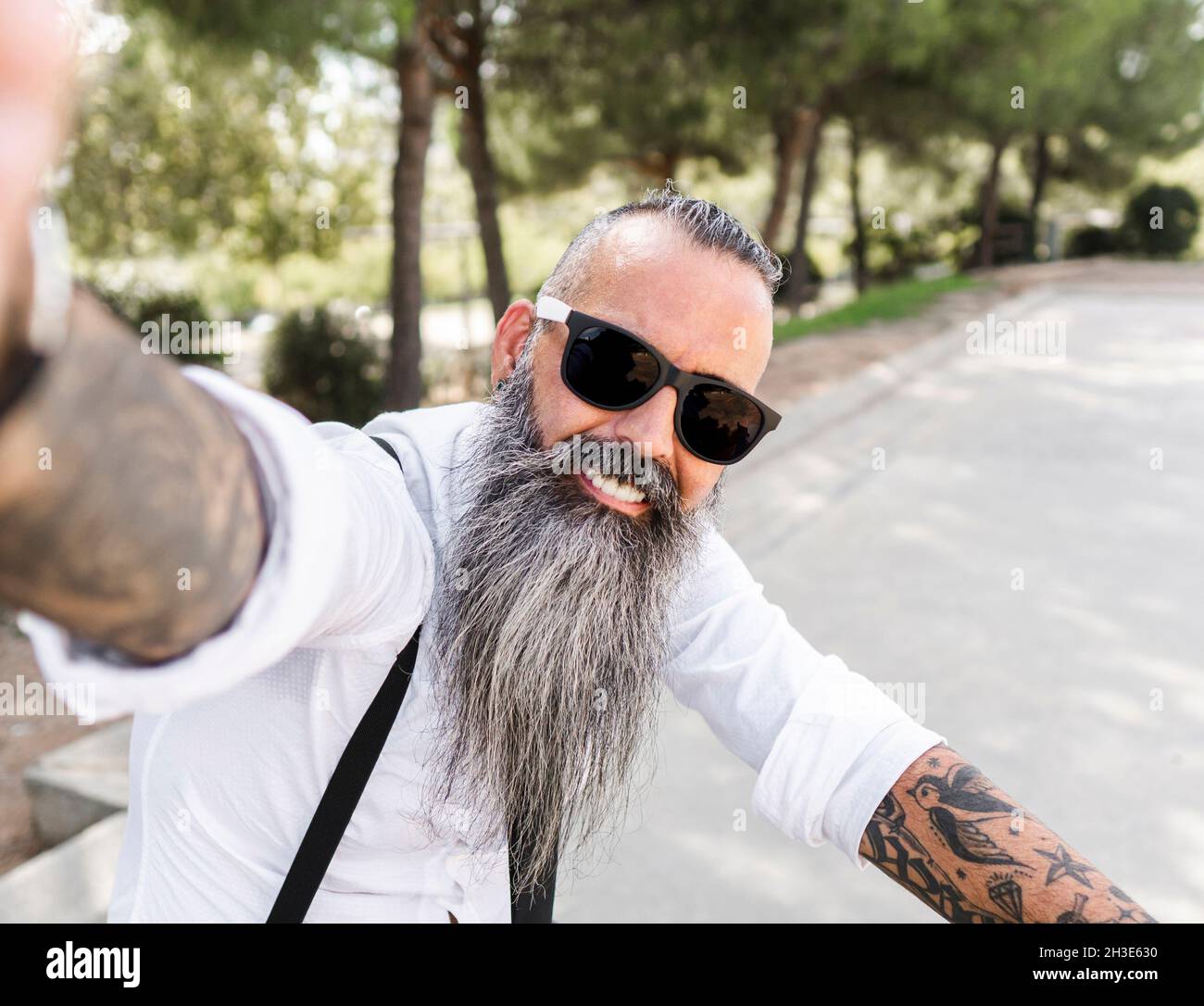 Positive bearded male in sunglasses and white shirt taking self portrait on smartphone while sitting on bicycle in park with trees Stock Photo
