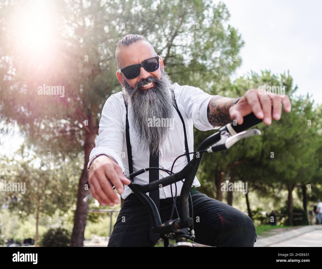 Confident happy male hipster with tattoos in white shirt and sunglasses sitting on bicycle in park with green trees in city Stock Photo