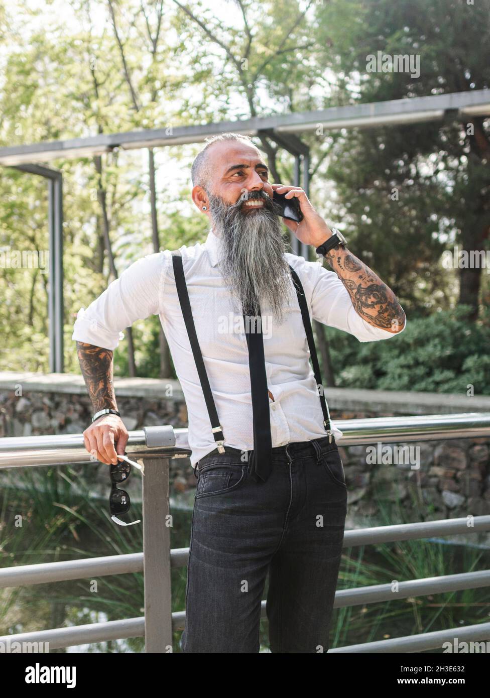 Confident smiling bearded hipster in stylish outfit speaking on cellphone while standing near metal fence on sunny street Stock Photo