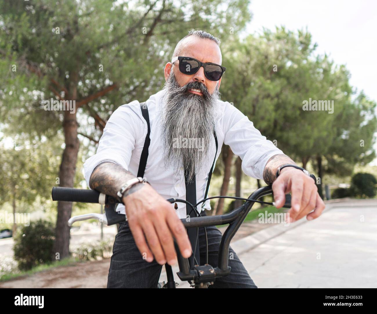 Confident male hipster with tattoos in white shirt and sunglasses sitting on bicycle in park with green trees in city Stock Photo