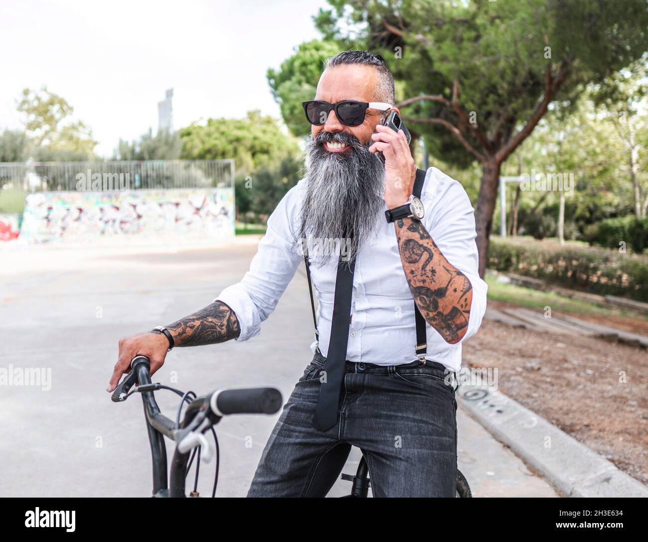 Bearded male in sunglasses and white shirt speaking on smartphone while sitting on bicycle in park with trees Stock Photo
