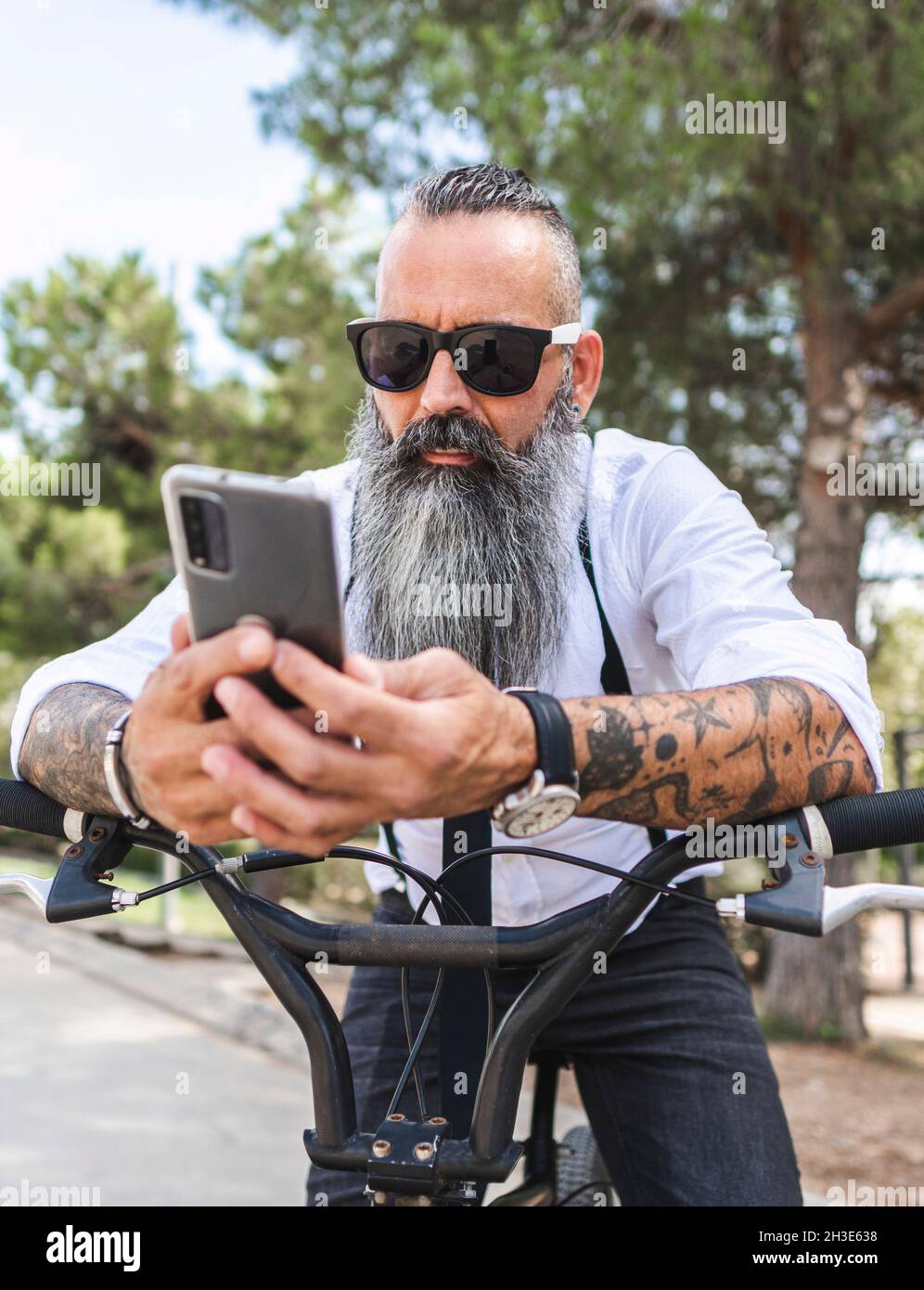 Bearded male in sunglasses and white shirt browsing on smartphone while sitting on bicycle in park with trees Stock Photo