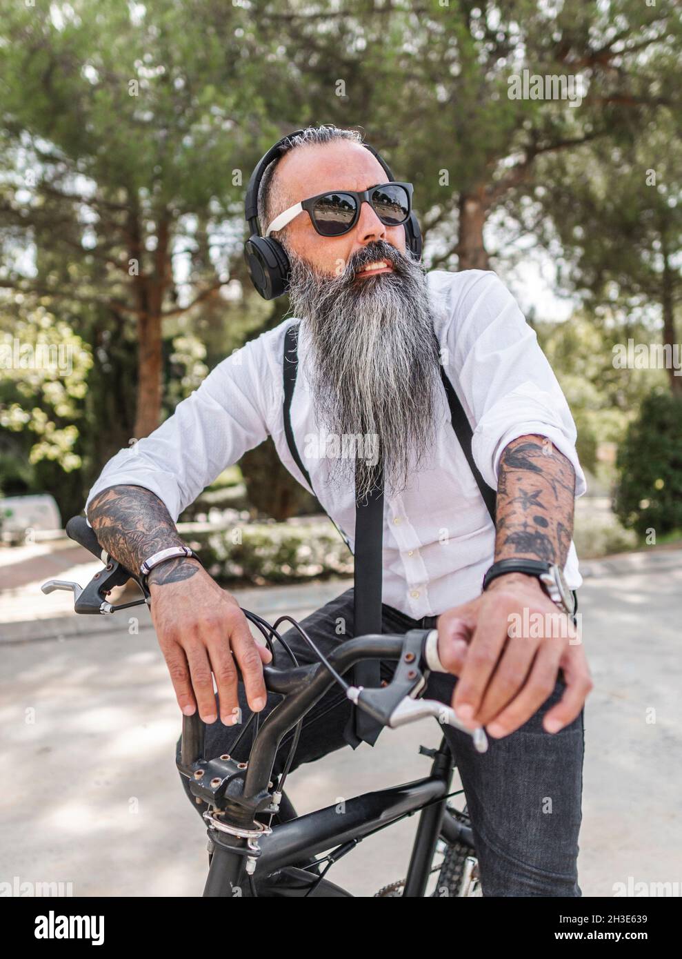 Confident male hipster in white shirt listening to music in headphones while sitting on bicycle on street with green trees Stock Photo