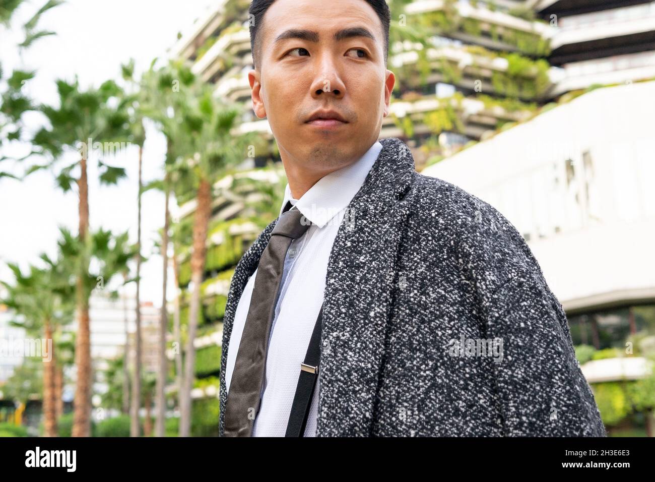 Well dressed young Asian male entrepreneur in tie looking away while strolling on road against modern buildings in city Stock Photo