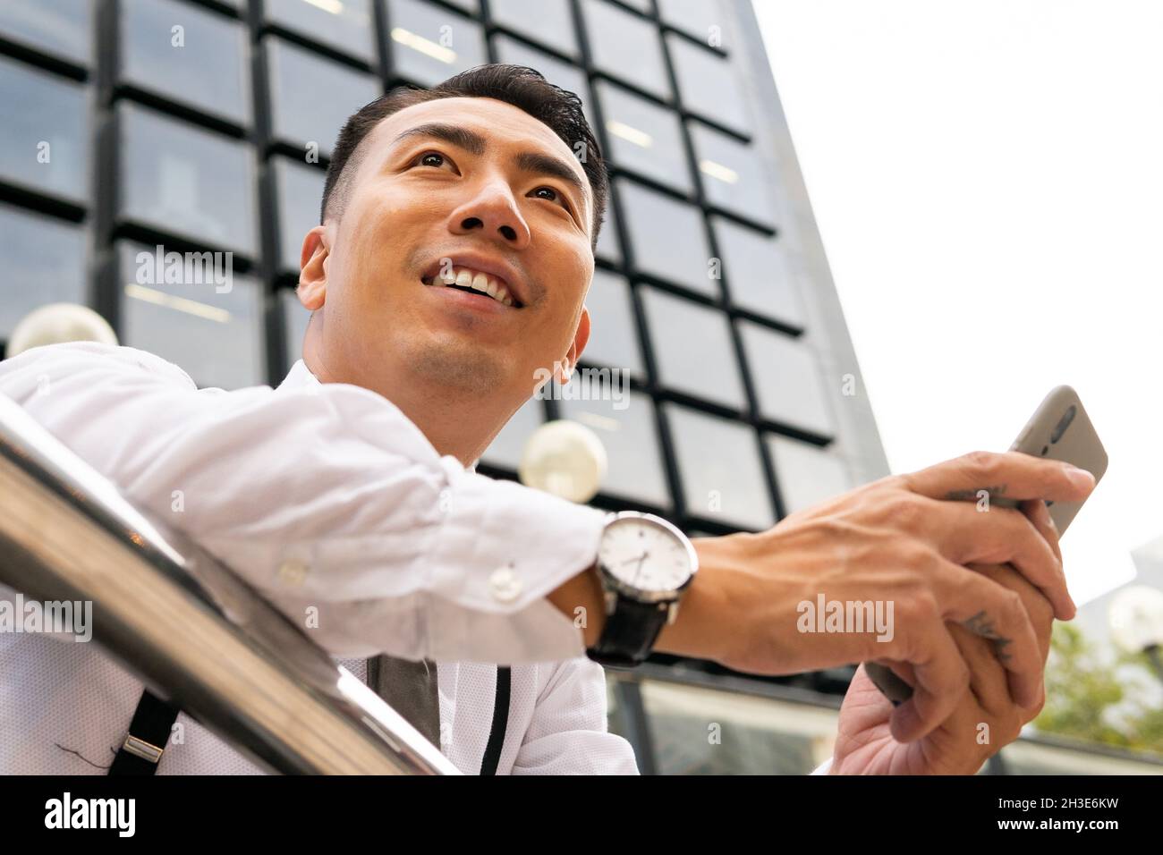 Low angle of well dressed young ethnic male entrepreneur in wristwatch text messaging on cellphone in city leaning on fence Stock Photo