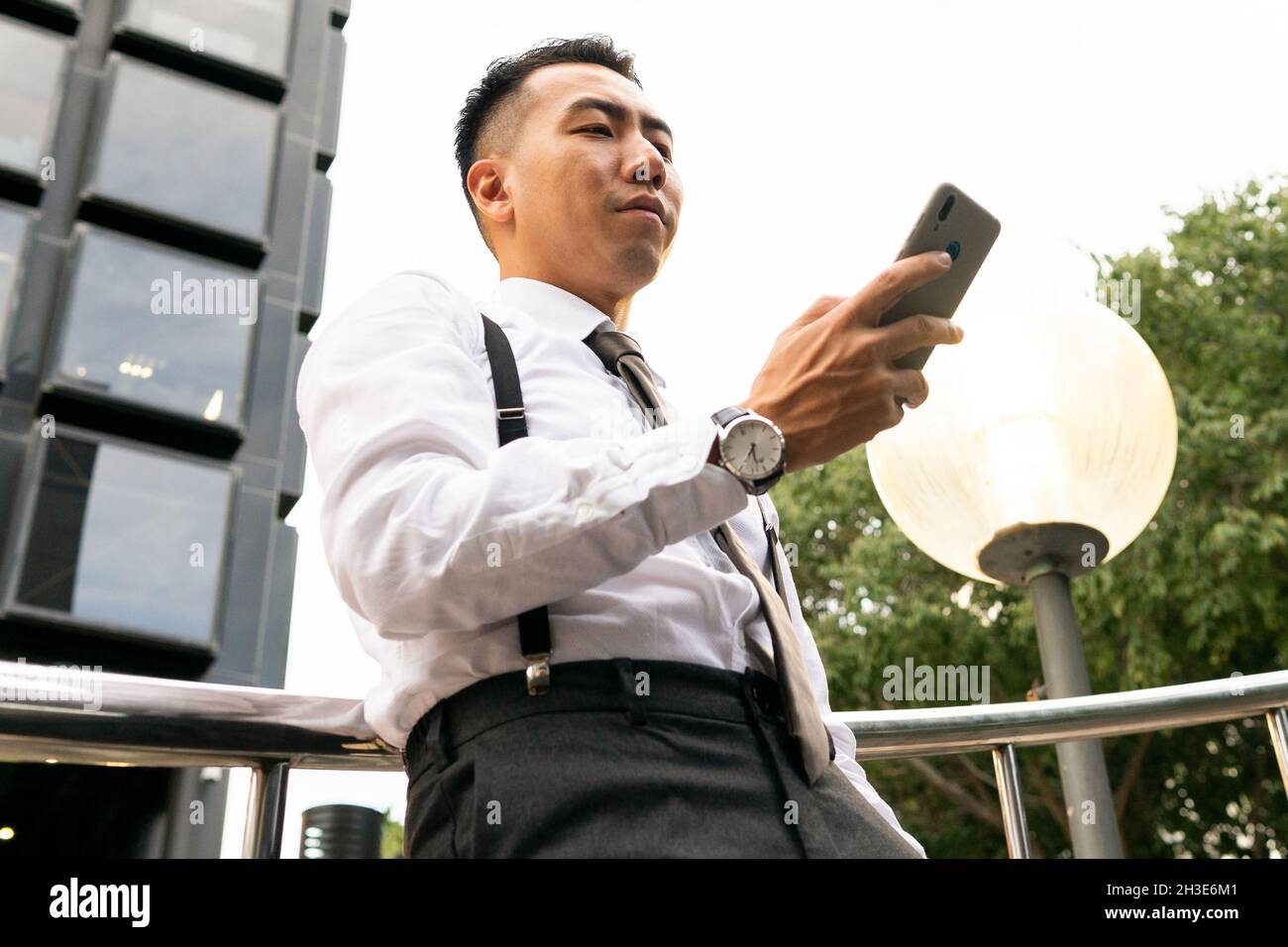 Low angle of well dressed young ethnic male entrepreneur in wristwatch text messaging on cellphone in city Stock Photo