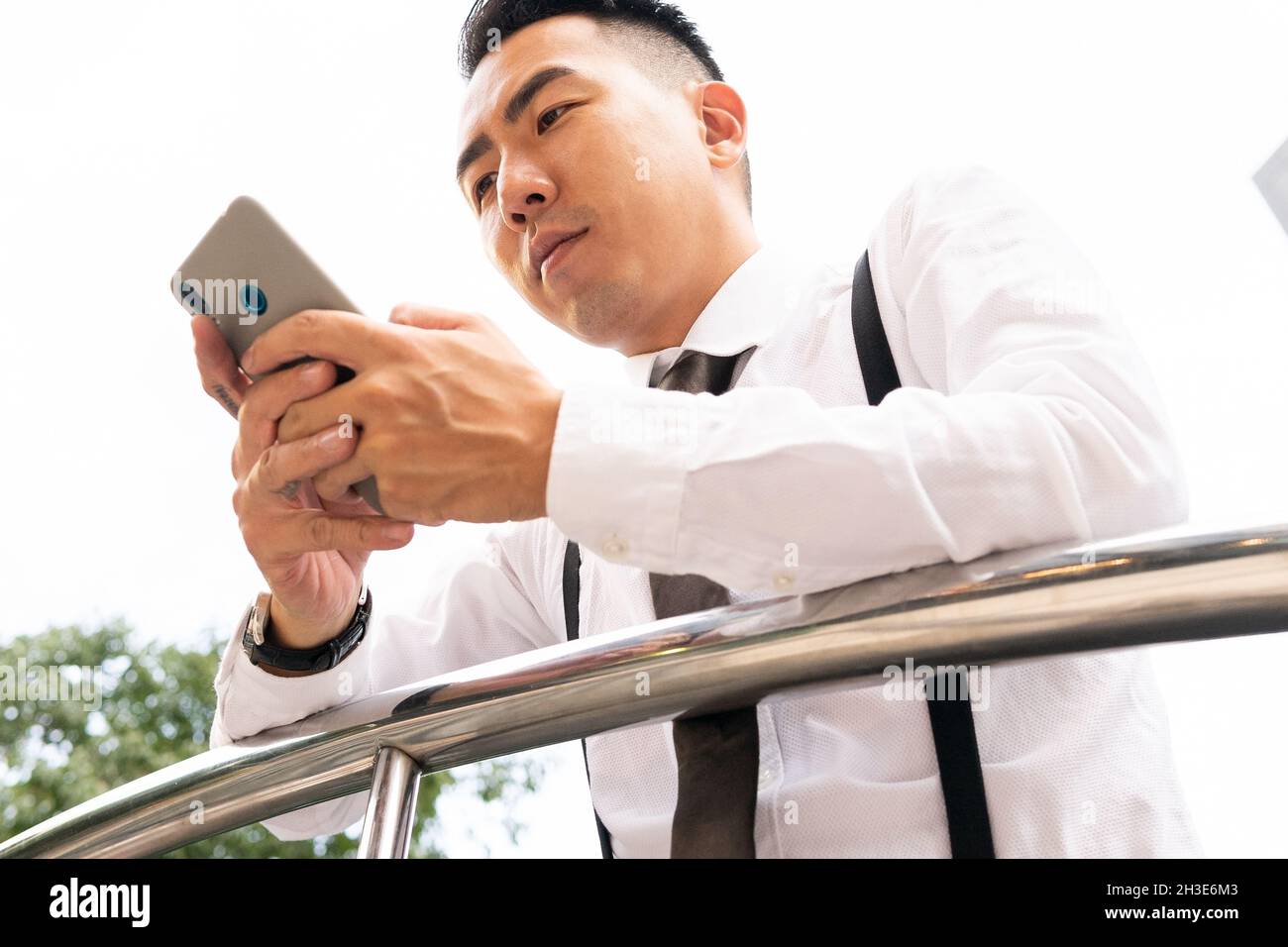 Low angle of well dressed young ethnic male entrepreneur in wristwatch text messaging on cellphone in city leaning on fence Stock Photo