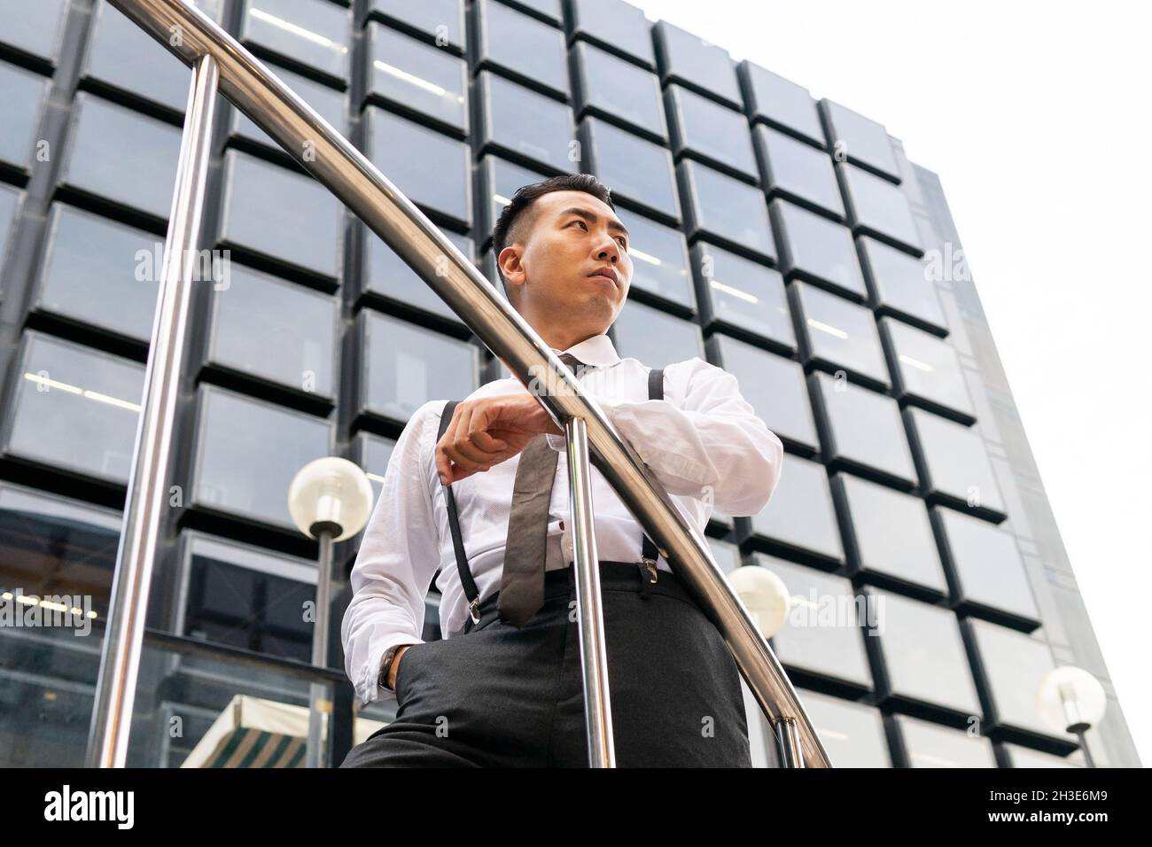 Low angle of well dressed young ethnic male entrepreneur with hand in pocket looking away between railing and multistory house in city Stock Photo