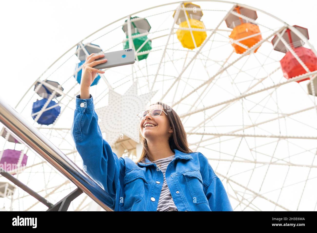 From below of smiling female taking self portrait while standing near colorful public Ferris wheel while standing in amusement park Stock Photo