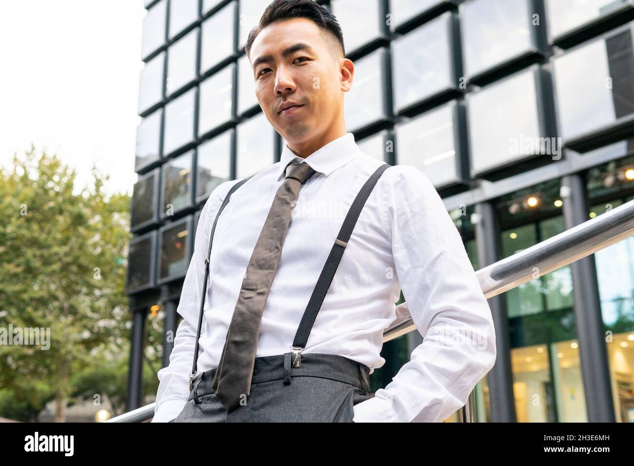 Well dressed young ethnic male entrepreneur with hand in pocket looking at camera between railing and multistory house in city Stock Photo