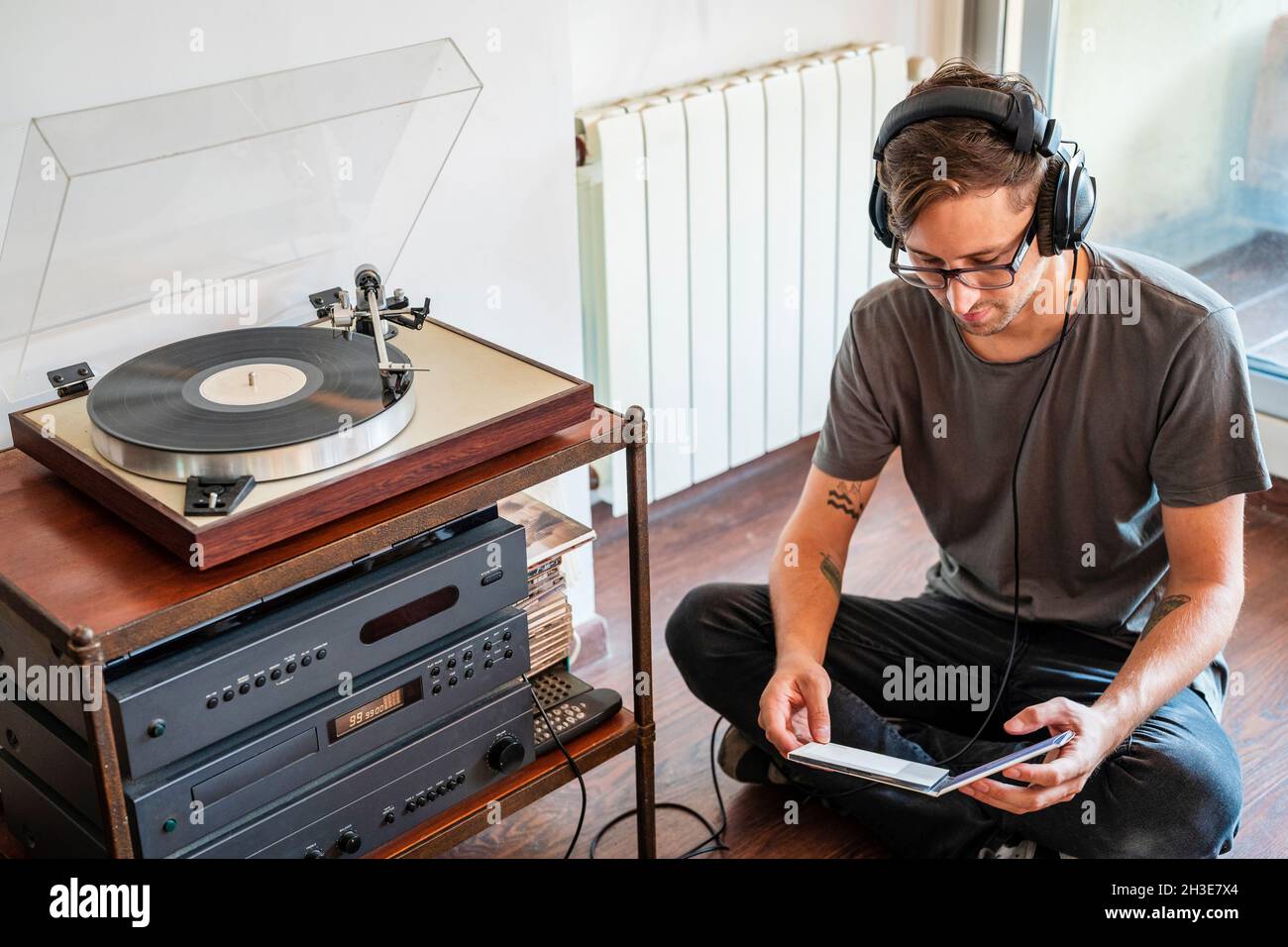 From above of man in casual clothes and headphones reading disc packaging while sitting on floor near record player Stock Photo
