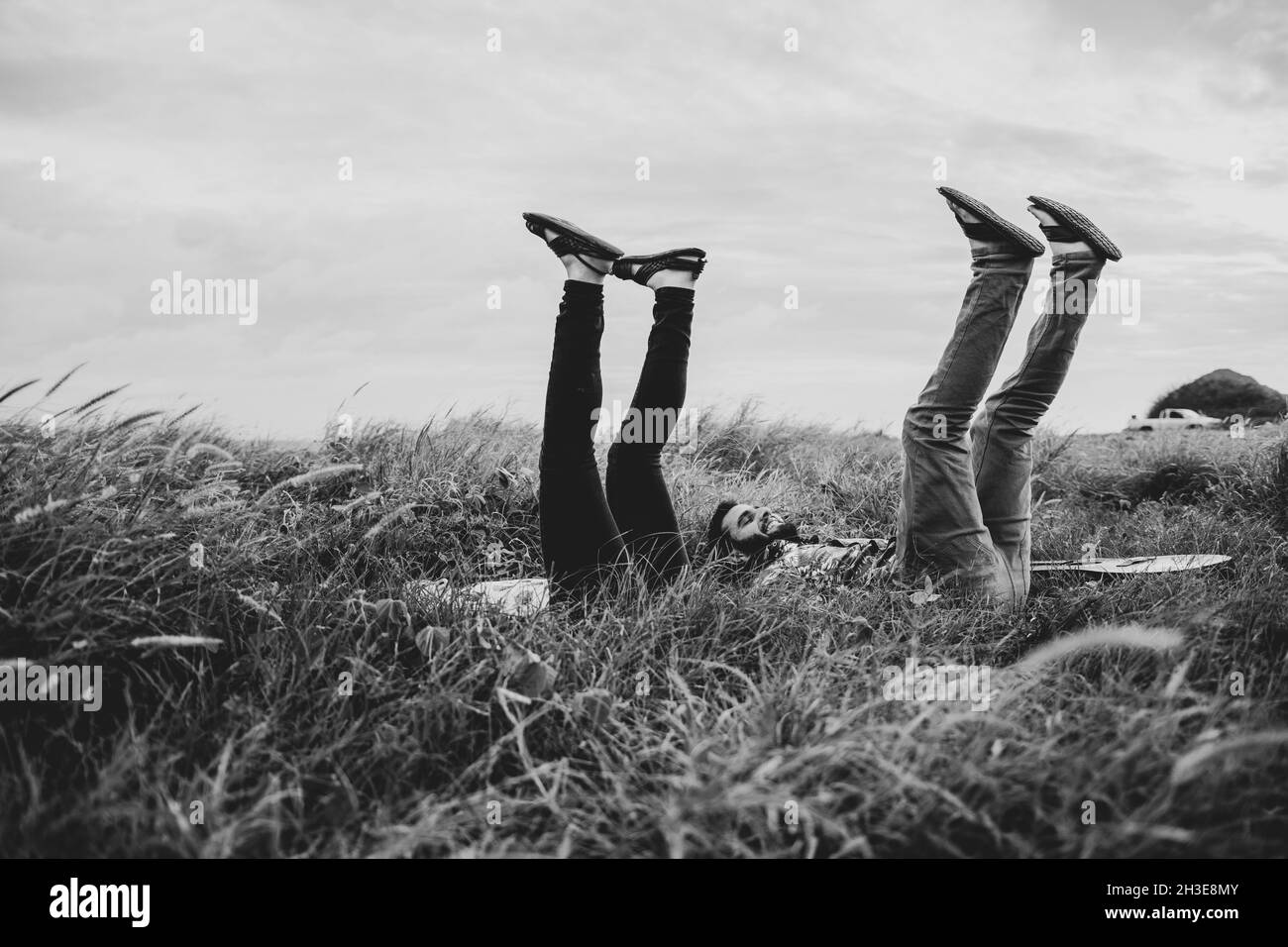 Black and white of side view of cheerful friends in casual clothes lying with raised legs on grassy field in nature in daylight Stock Photo