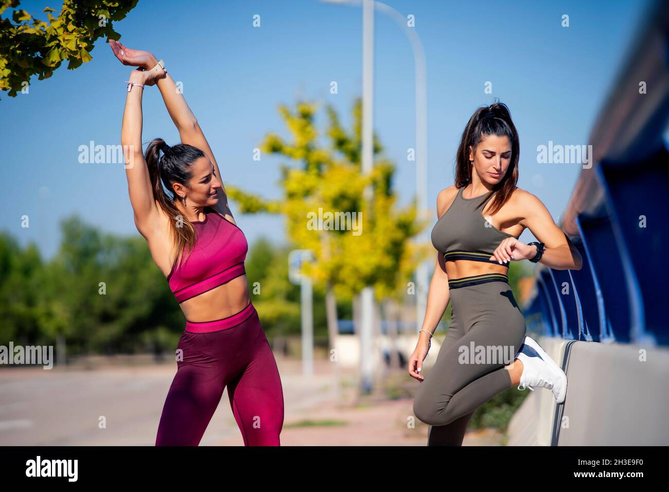 Slim lady in sportswear stretching arms while standing on sports ground near twin sister looking at fitness bracelet Stock Photo