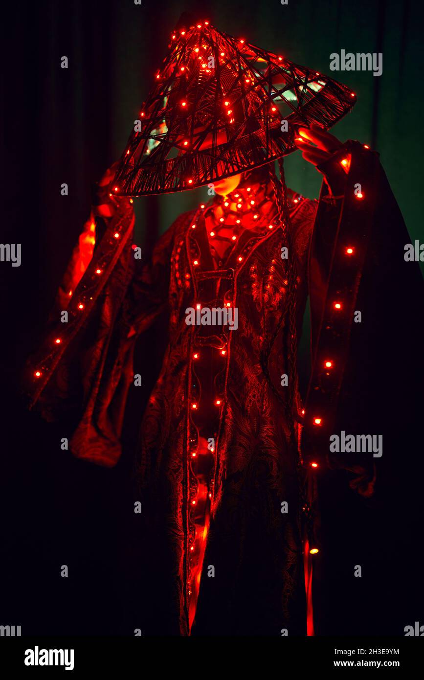Unrecognizable enigmatic female in creative traditional outfit and Vietnamese headwear with red illumination standing in dark studio on black backgrou Stock Photo