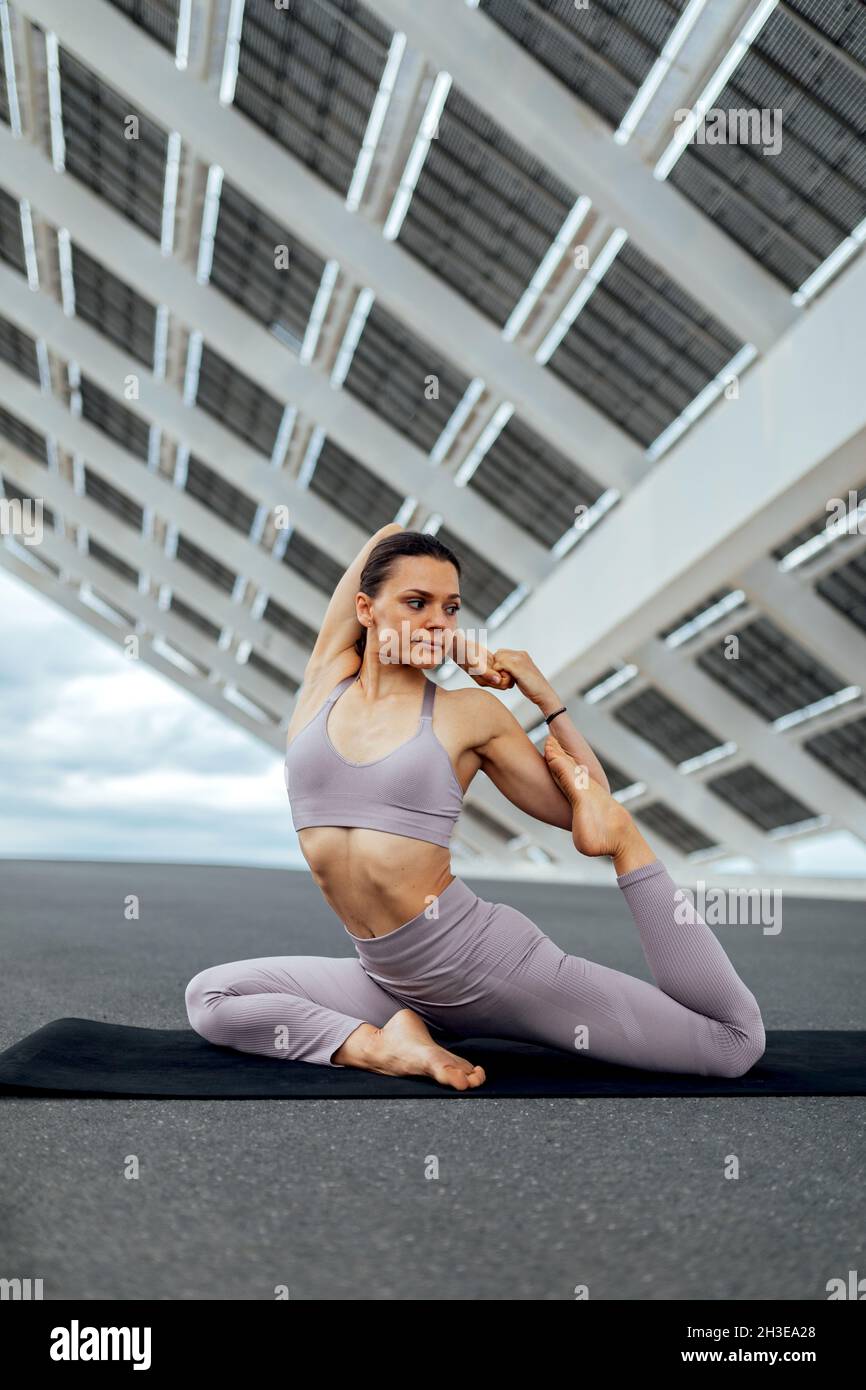 Full body of female doing Eka Pada Raja Kapotasana posture while sitting on mat on street near solar panel during yoga training Stock Photo