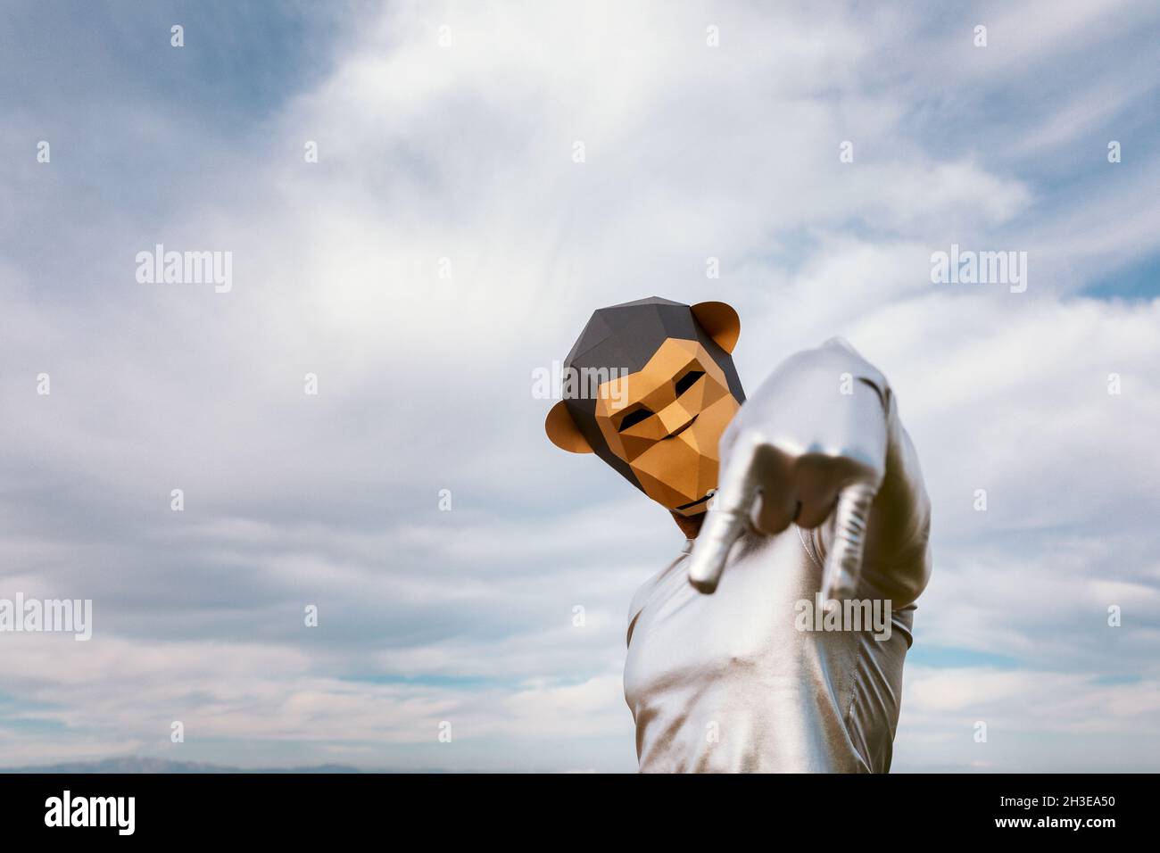 Self assured person with geometric monkey mask showing index finger and pinky down against clouds Stock Photo