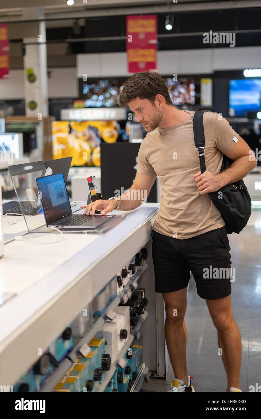 Casual guy in shorts and t shirt looking at screen of modern laptop while choosing new device in electronic store Stock Photo