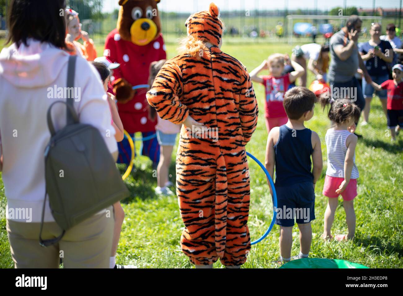 A teenager in a tiger costume stands in the park, view from behind. Crowd of people, celebration in the fresh air. Sunny day. City Lifestyle Stock Photo