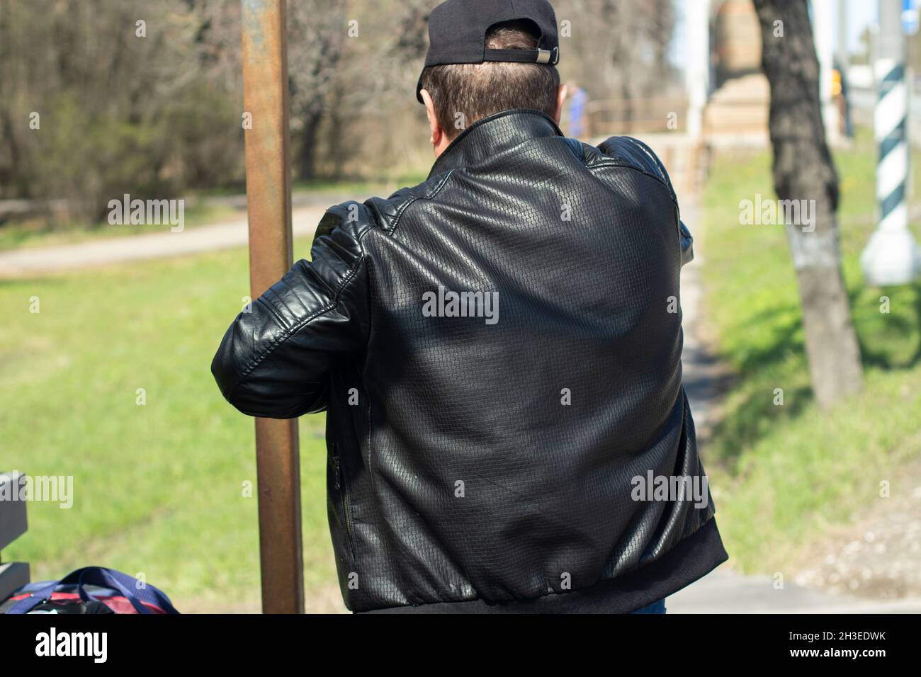 A man in a black leather jacket and cap is waiting for the bus, a view from the back. A day out of town. Traffic. Late autumn Stock Photo